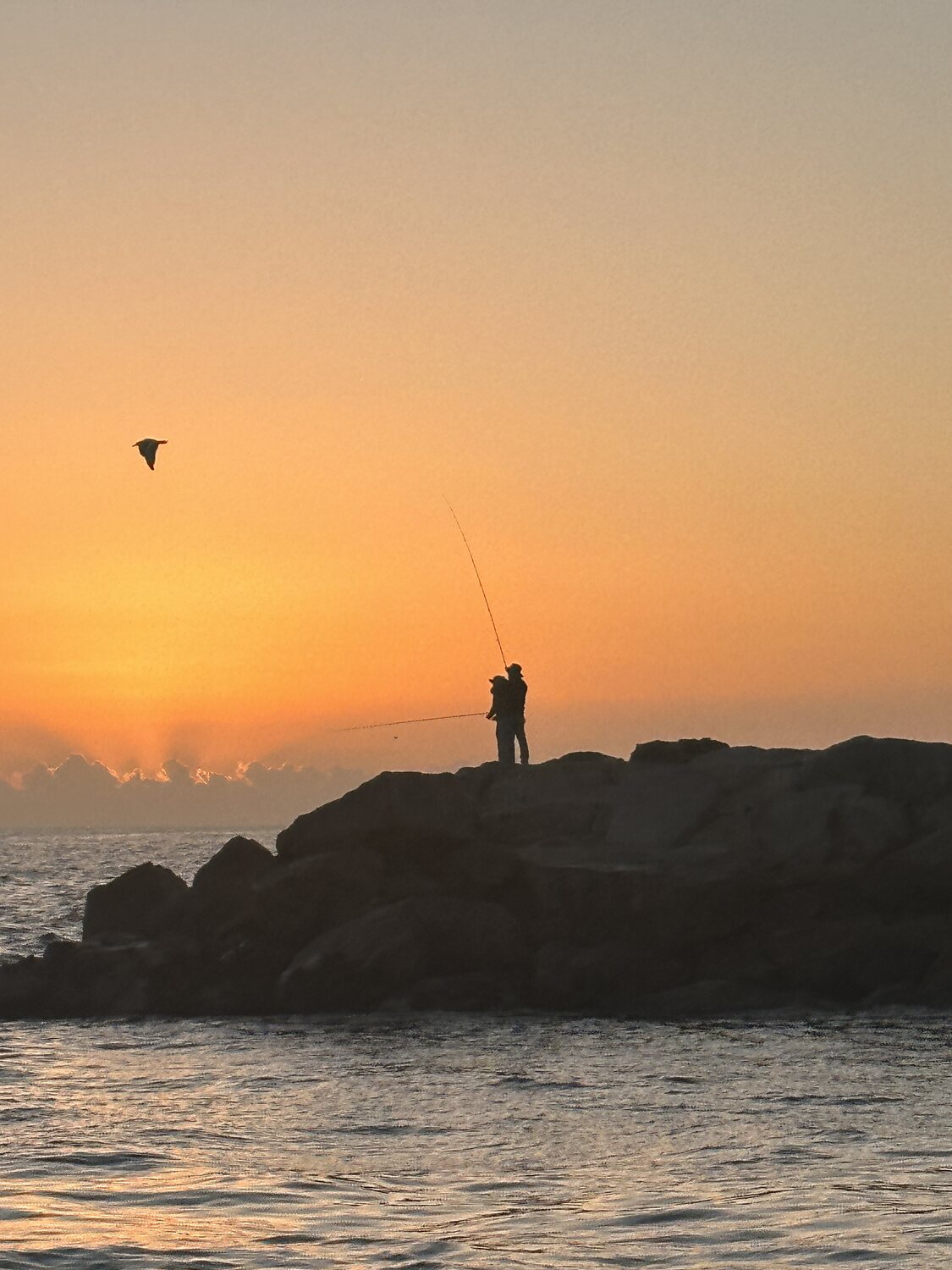 Two figures fishing in silhouette on a rock jetty at sunrise, a gull crossing the orange sky